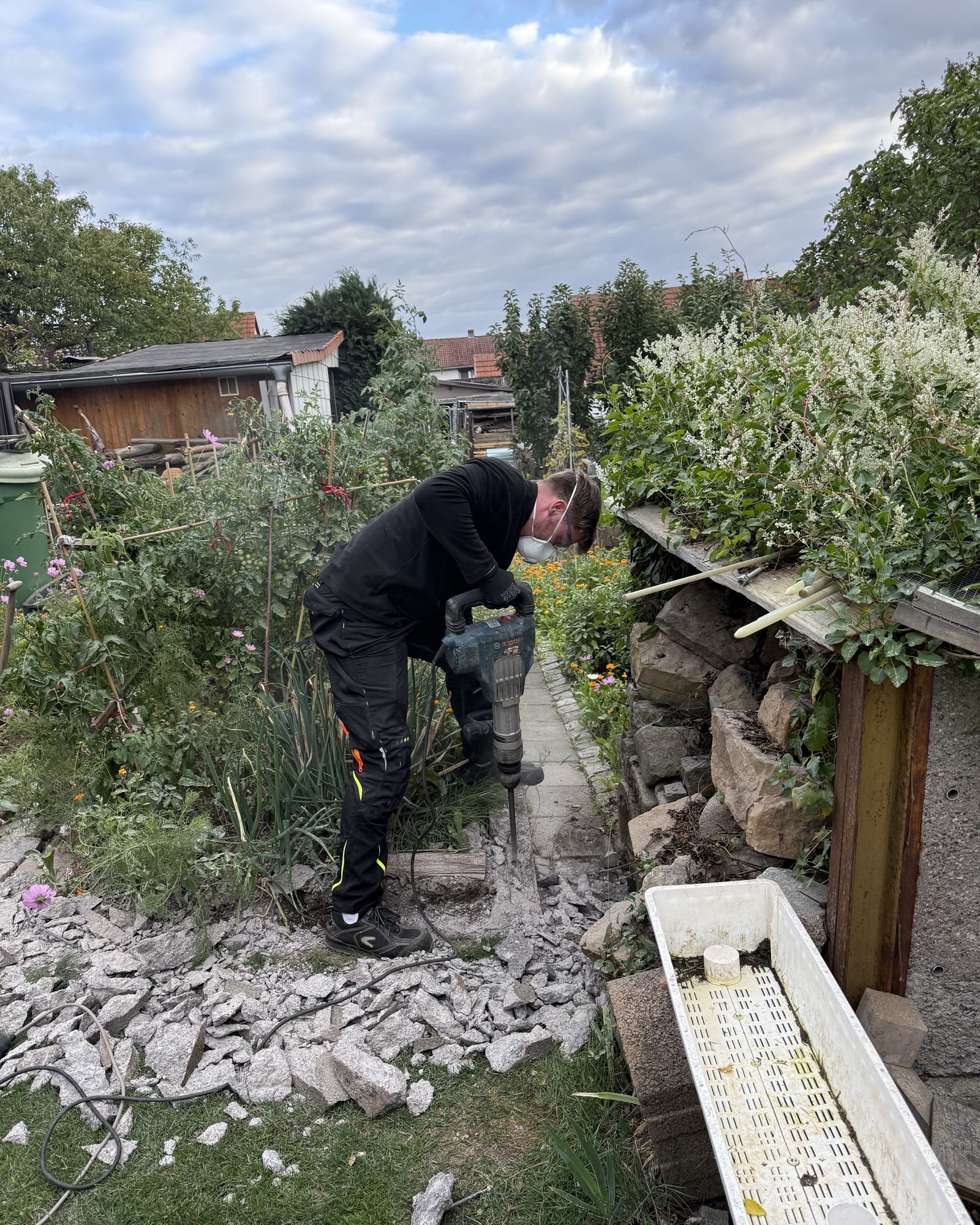Einer von uns bricht mit Staubschutzmaske und Abbruchhammer alten Betonbelag in einem Garten auf